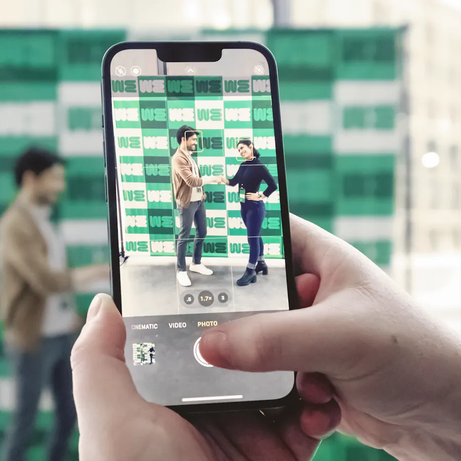 Two hands holding a phone taking a picture of two people shaking hands in front of a backdrop banner.