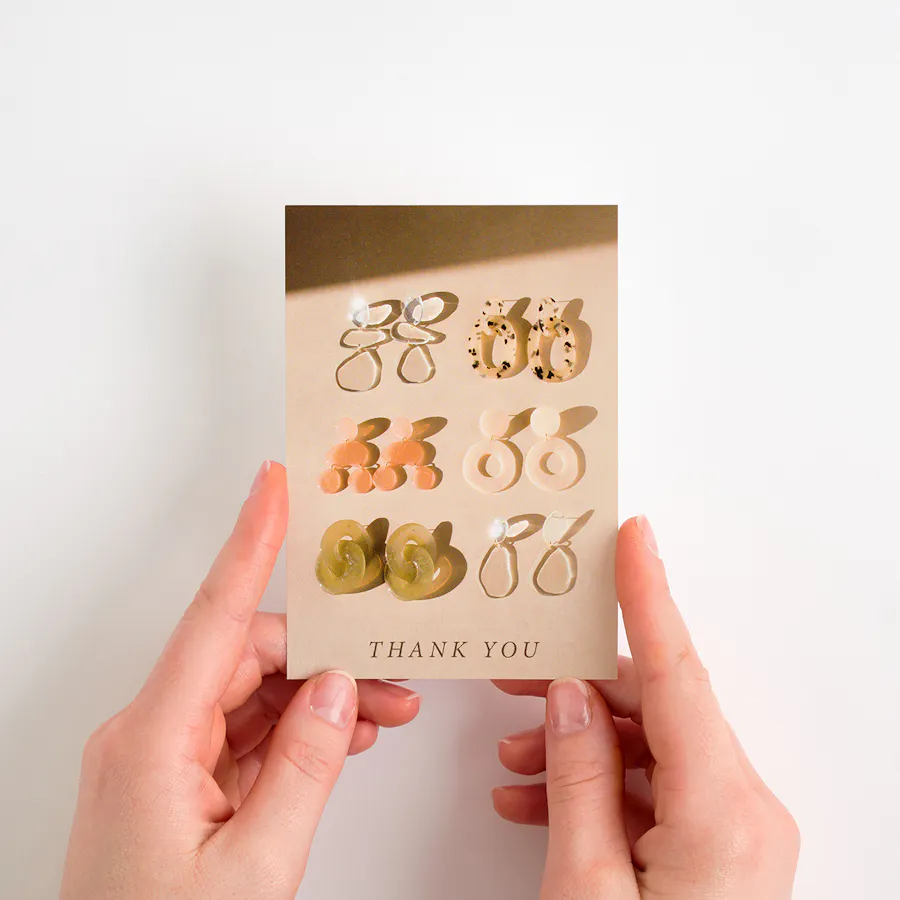 Two hands holding a postcard flyer printed with six pairs of earrings and Thank You at the bottom.