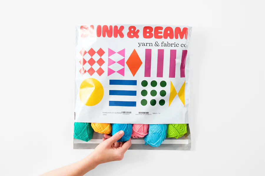 A hand pulling a skein of yarn out of a custom poly mailer printed with Blink & Beam and multicolored graphics.