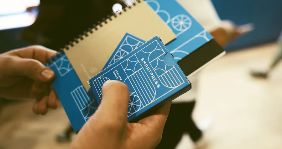 Two hands holding custom printed playing cards and a notebook with a blue and white Smartpress design.