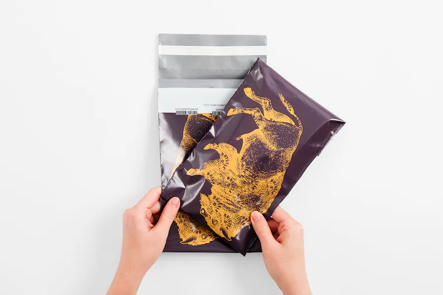 Two hands holding two mailer shipping bags printed with a dark purple background and golden yellow buffalo graphic.