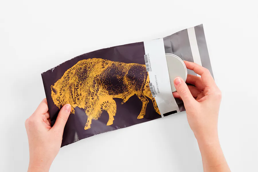 Two hands holding a poly mailer bag printed with a buffalo graphic and pulling a silver container from the bag.