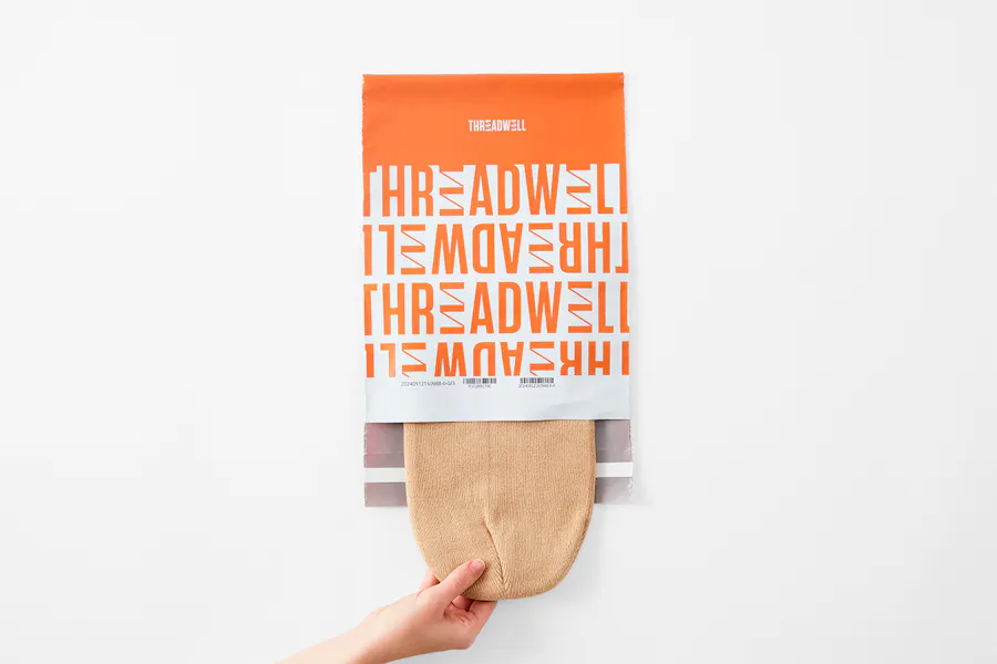 A hand pulling a knit beanie out of a custom poly mailer printed with an orange and white Threadwell design.