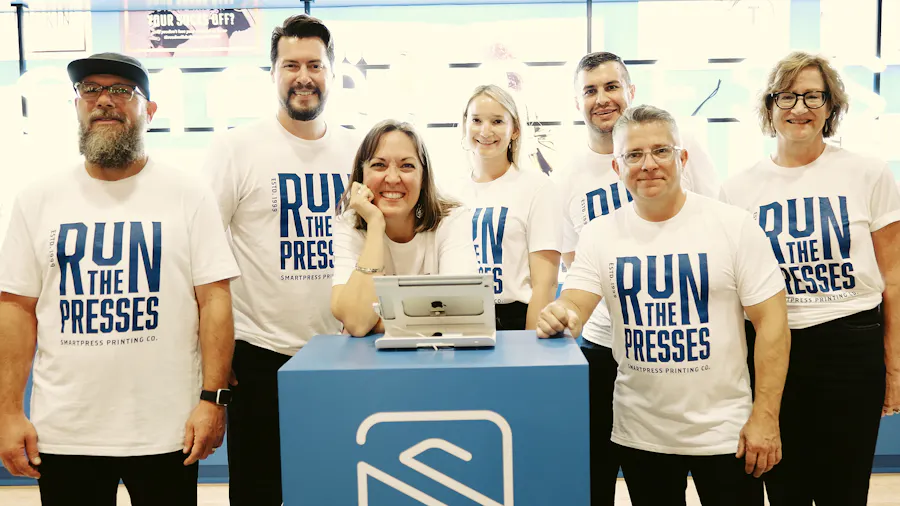 Seven smiling people in front of the Smartpress Adobe MAX booth wearing white shirts printed with Run the Presses.