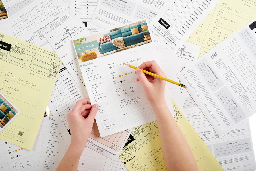 A hand holding a carbonless form order receipt and another hand writing on it with a pencil above a pile of more carbonless forms.