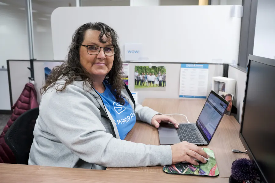 A woman wearing a blue Smartpress T-shirt, sitting at a desk in a cubicle and working on a laptop.