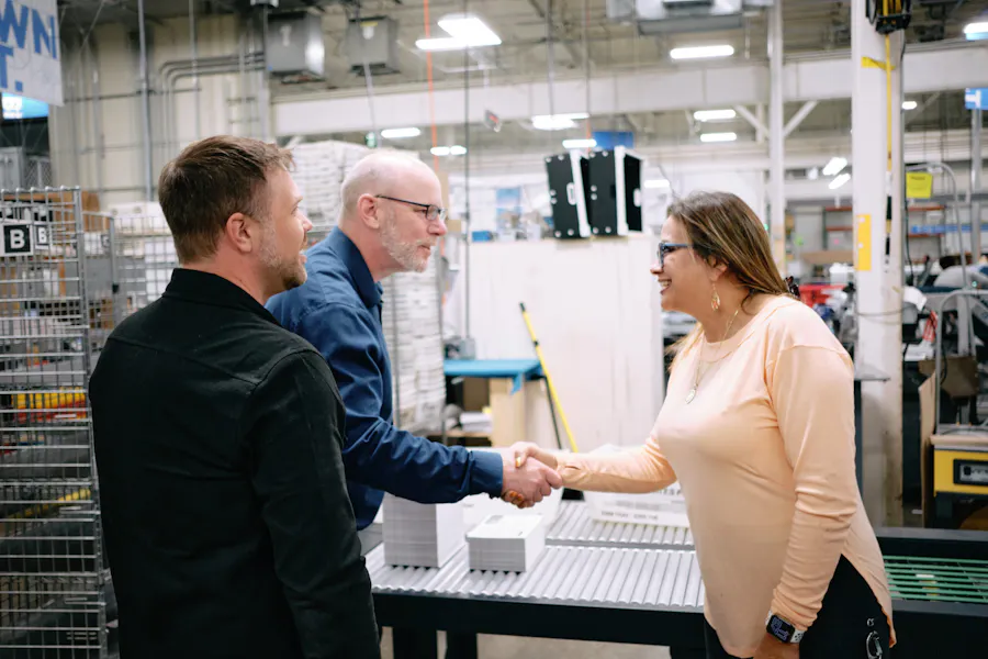 Three people shaking hands and talking in a print production facility with machinery all around them.