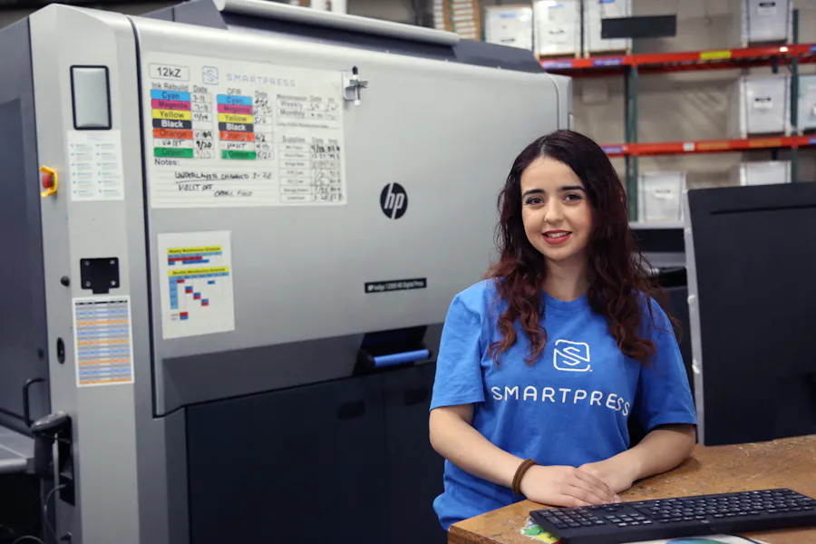 A smiling woman standing at a desk with a computer on it in a print production facility.