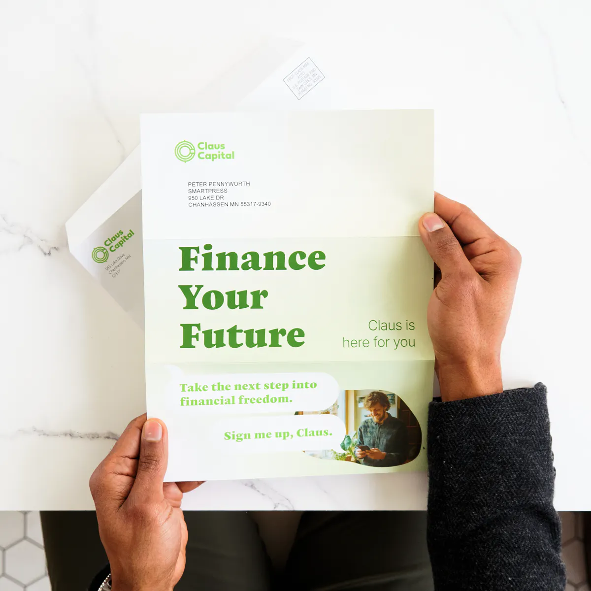 Two hands holding a letter mailer with finance information and a matching envelope underneath it.