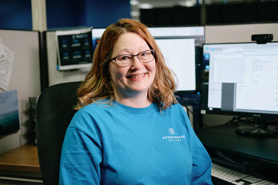 A smiling woman wearing a blue Smartpress shirt sitting at her desk with two computer monitors on it.