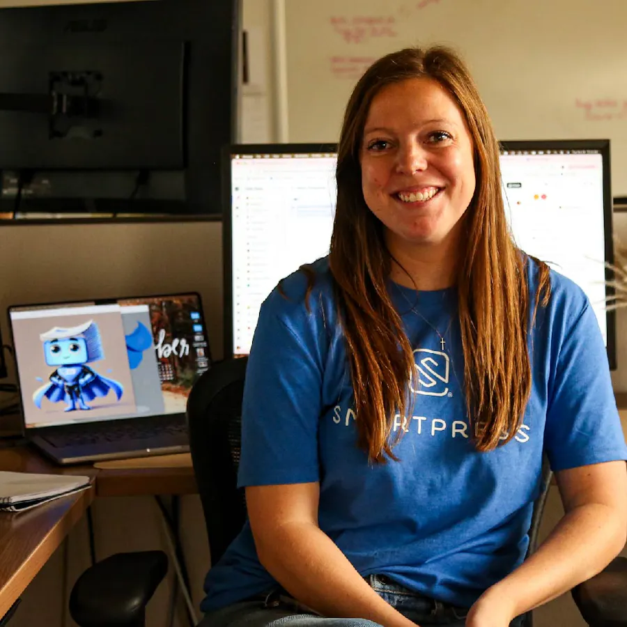A smiling woman with dark red hair wearing a blue Smartpress T-shirt and sitting at a desk in an office.