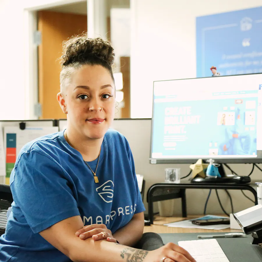 A woman wearing a blue Smartpress T-shirt and sitting at a desk in a cubicle in an office.