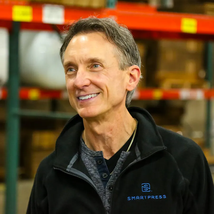 A smiling man in a production facility wearing a black Smartpress zipup jacket.