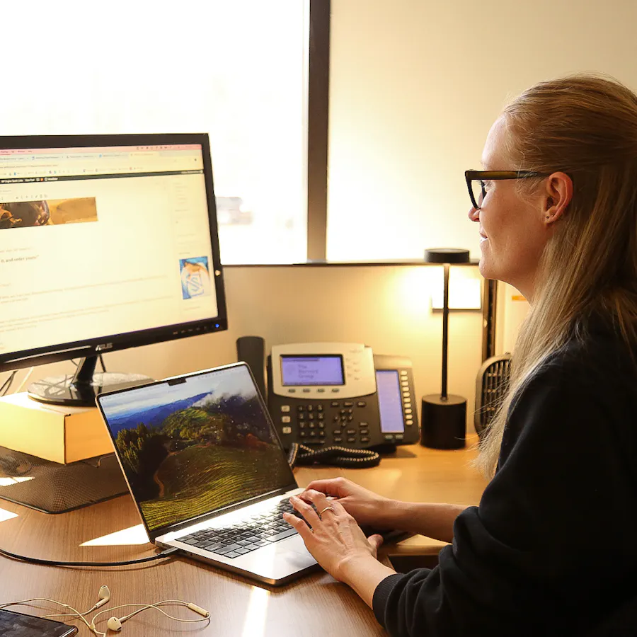 A woman sitting at a computer desk and typing on a laptop while wearing a black hoodie.
