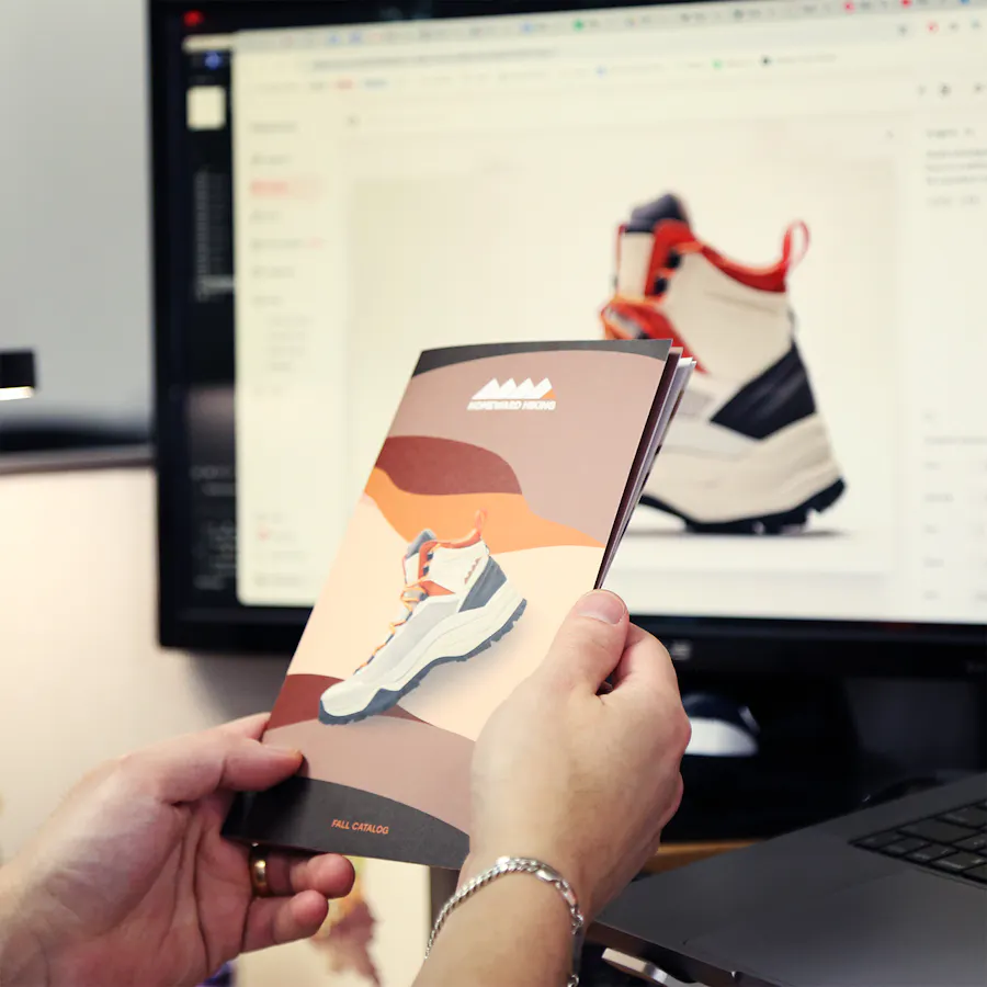 Two hands holding a custom booklet printed with a sneaker image in front of a desktop screen showing the same thing.
