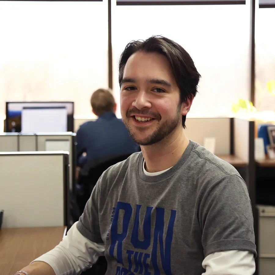 A smiling man with dark hair sitting at a desk in an office and wearing a gray T-shirt.