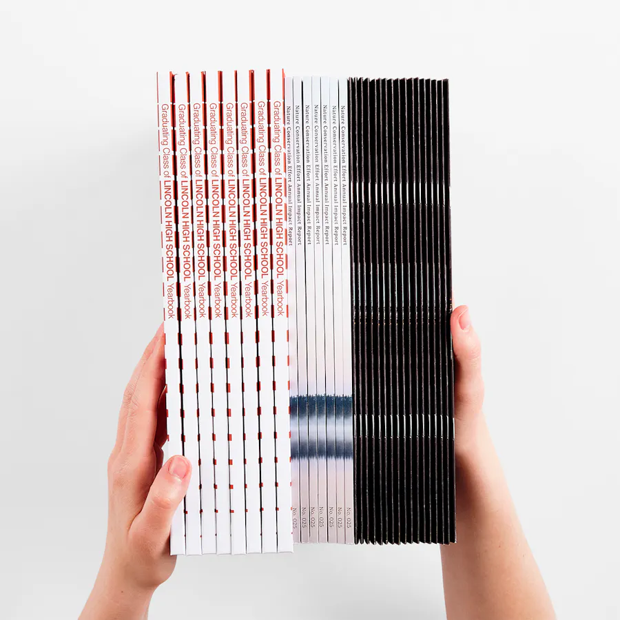 Two hands holding a stack of yearbooks with hardcover, perfect bound and saddle stitch bindings.