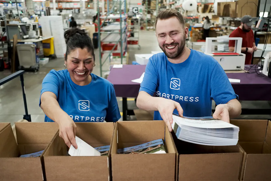A smiling woman and man in a Smartpress production facility putting booklets into boxes.