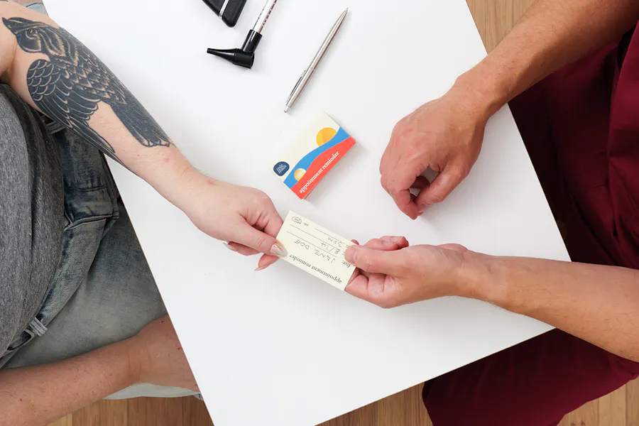 A nurse handing an appointment card to a patient while sitting at a white desk.