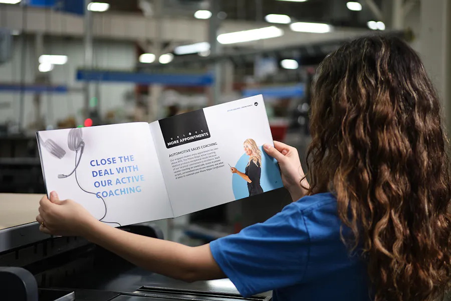 A woman in a print production facility holding open a marketing booklet about sales coaching.
