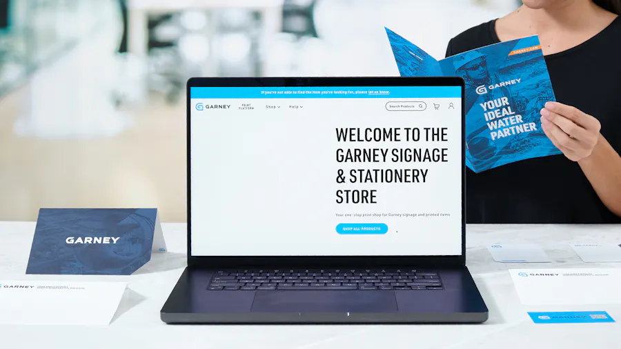 A laptop showing Garney's print management software with a person holding a brochure behind it.
