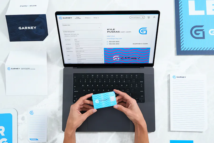Two hands holding a Garney business card over a laptop with more Garney print assets around it.