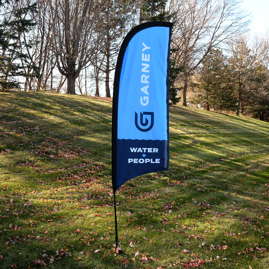 A Garney branded feather flag printed with shades of blue and installed on a grassy hill.
