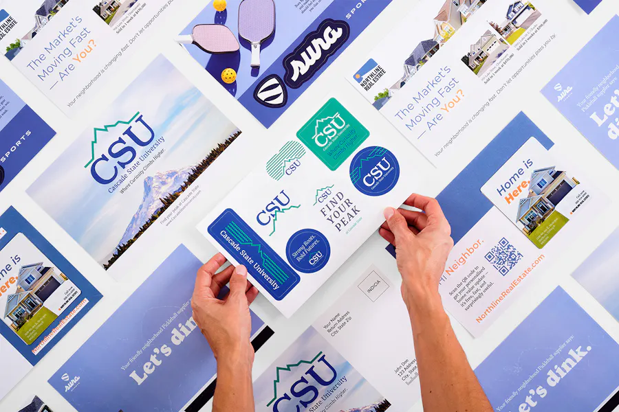 Two hands holding a sticker postcard with CSU branding and more postcards around it.