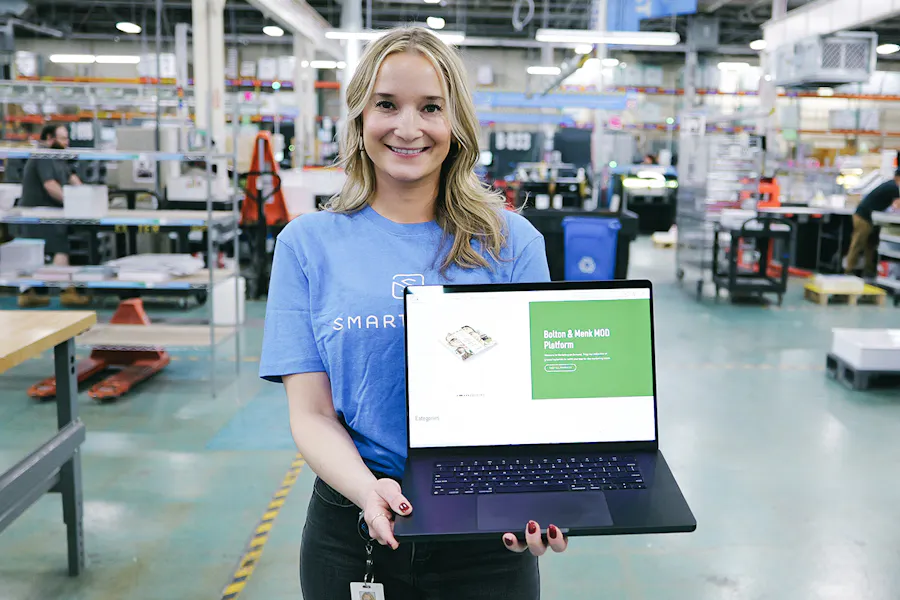 A smiling woman standing on a print production floor holding a laptop showing print management software.