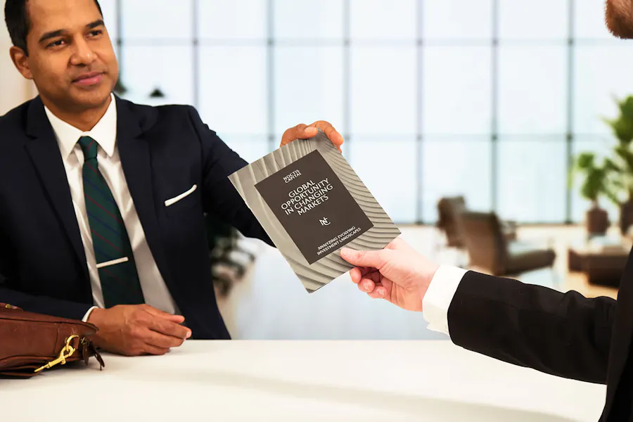 A hand passing a finance marketing white paper to a man wearing a suit sitting at a table.