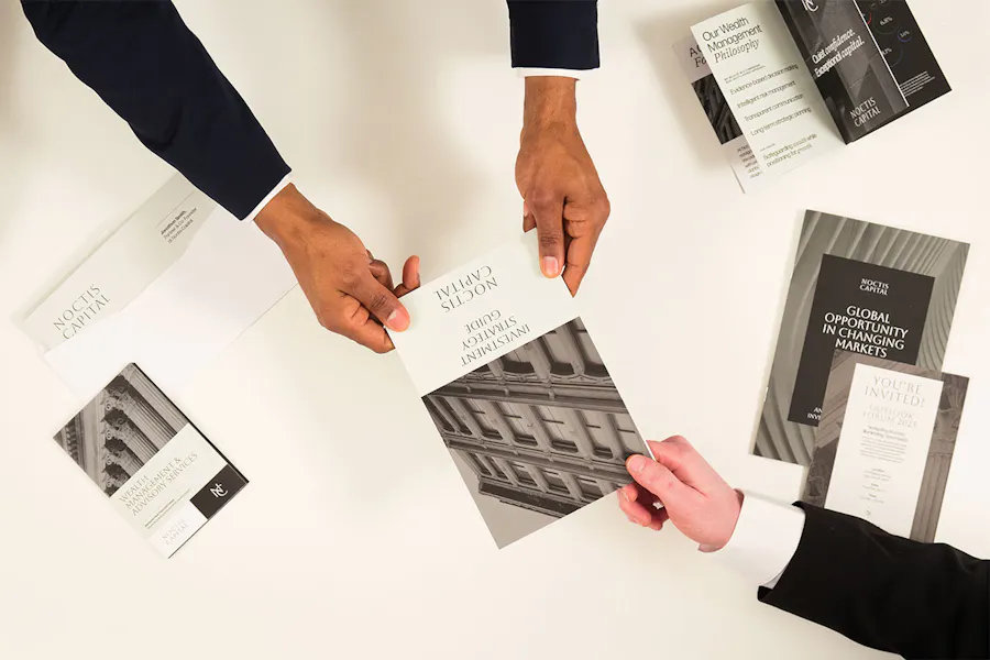 Three hands holding a finance marketing investment guide with other assets around them.