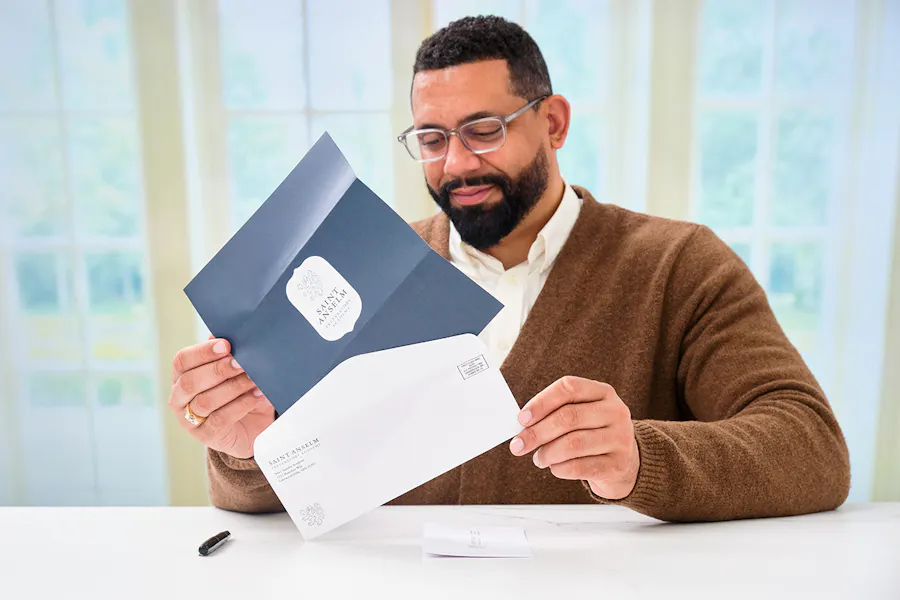 A man holding a letter mail and matching envelope printed with Saint Anselm branding in blue.