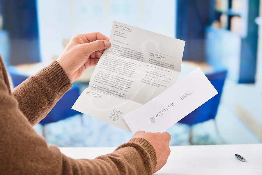 A person in a tan sweater holding a letter mailer and envelope printed with Saint Anselm information.