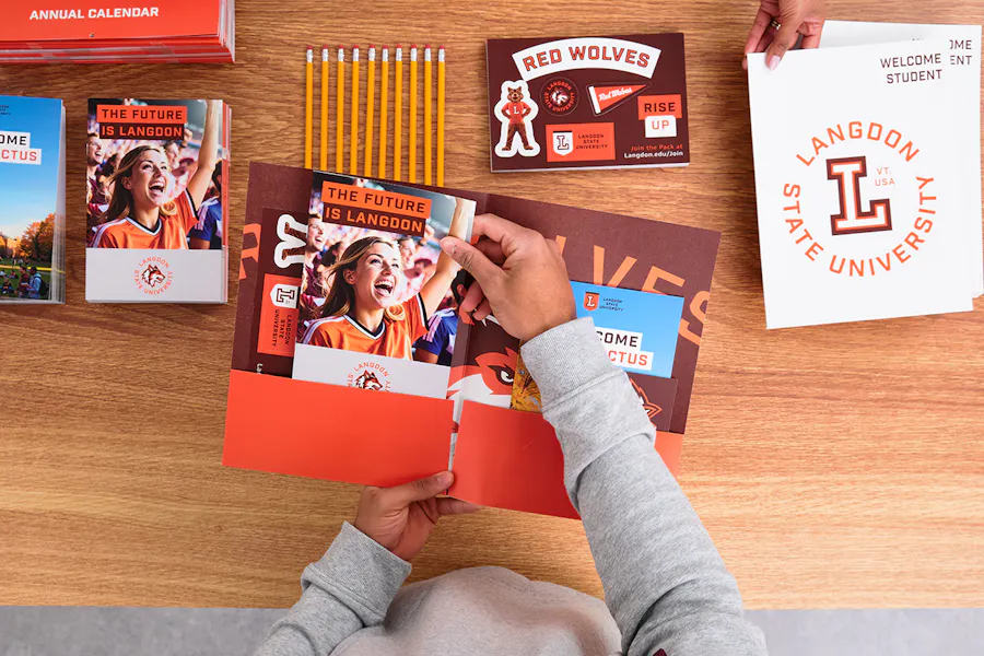 Two hands holding print marketing for schools in a pocket folder with other print pieces around them.