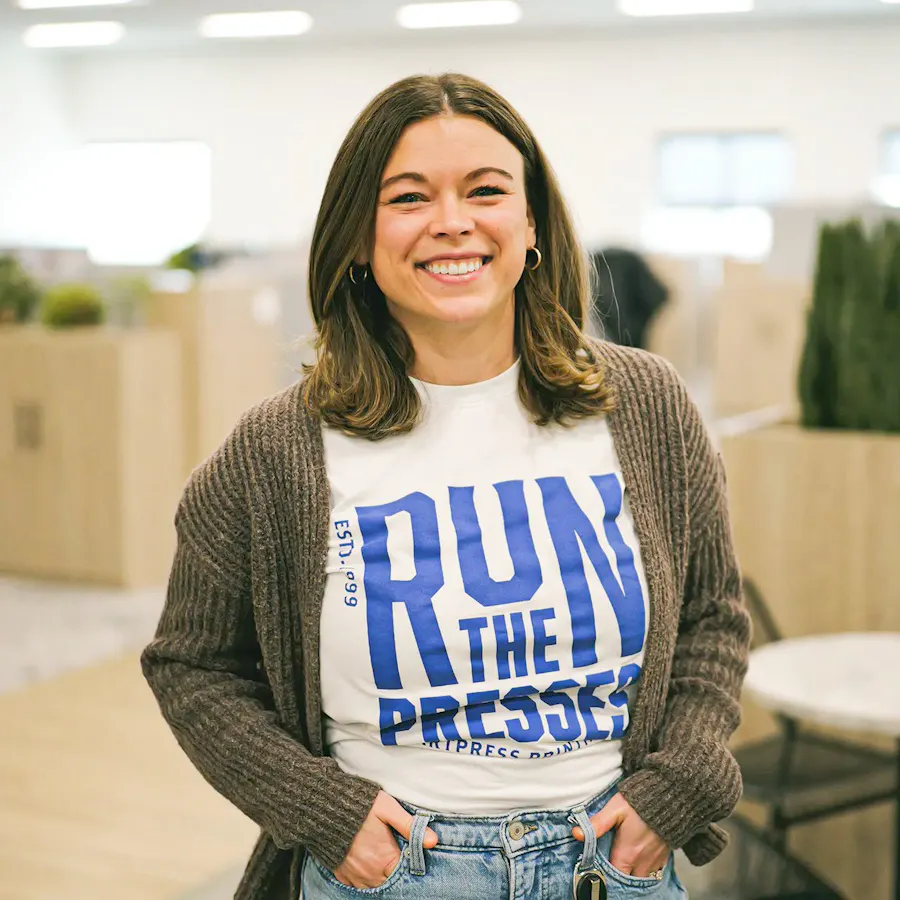 A smiling woman with brown hair standing in an office and wearing a tee with Run the Presses printed on it.