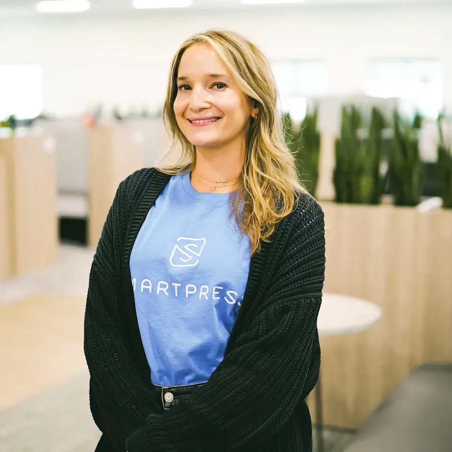 A smiling woman with blonde hair standing in an office and wearing a blue tee with Smartpress on it.