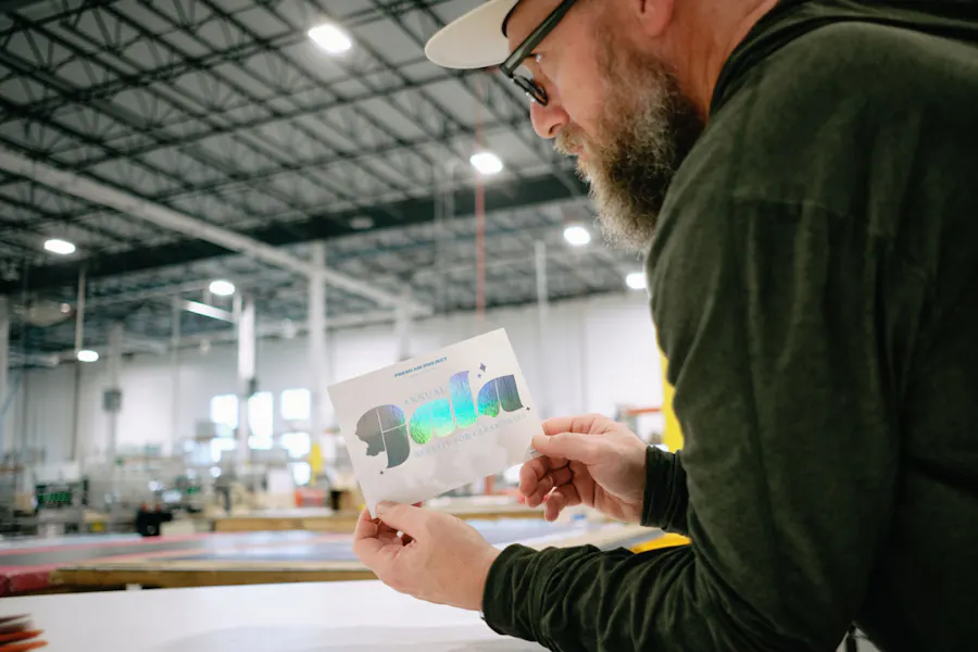 A man with glasses leaning on a white table in a production facility holding a printed invitation.