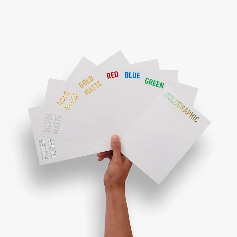 A hand holding eight pieces of white cardstock printed with eight different raised foil colors.