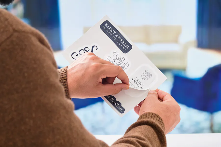 Two hands holding a sticker postcard printed with Saint Anselm branding.