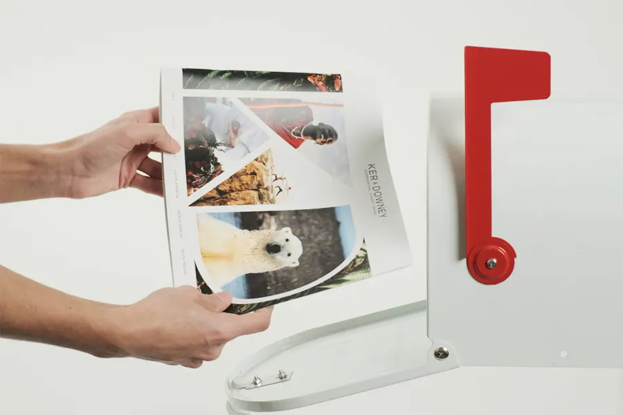 Two hands holding a printed booklet in front of an open mailbox with a red flag.