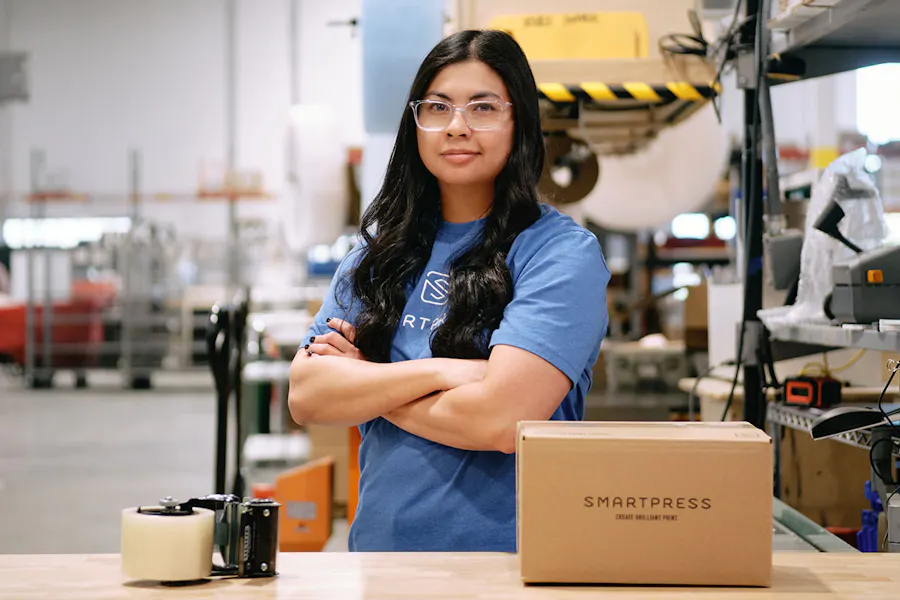 A woman in a blue Smartpress shirt standing by a table with a box on it in a production facility.